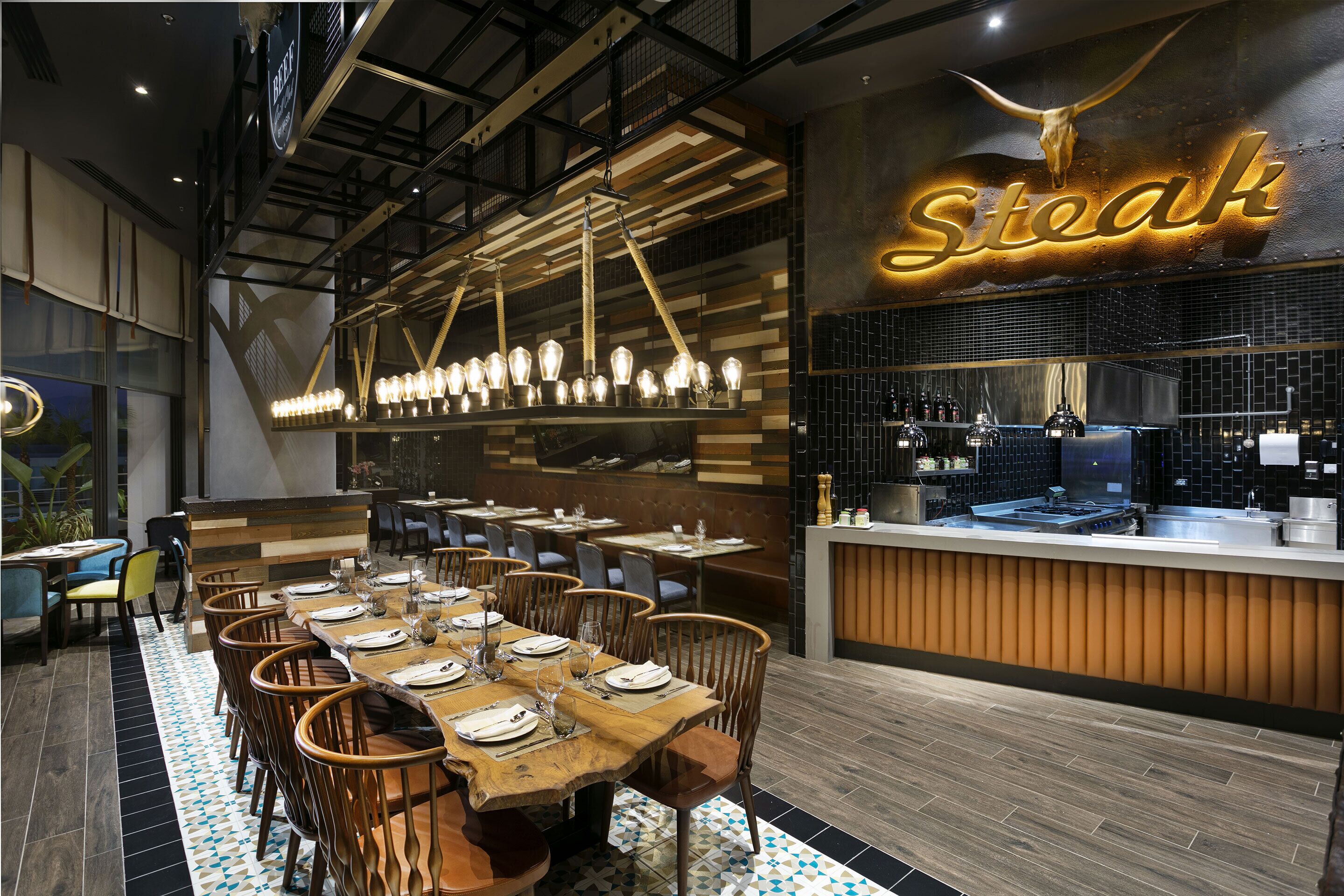 A restaurant interior with a long wooden table set for dining, a hanging light fixture, and a 'Steak' sign above the counter.