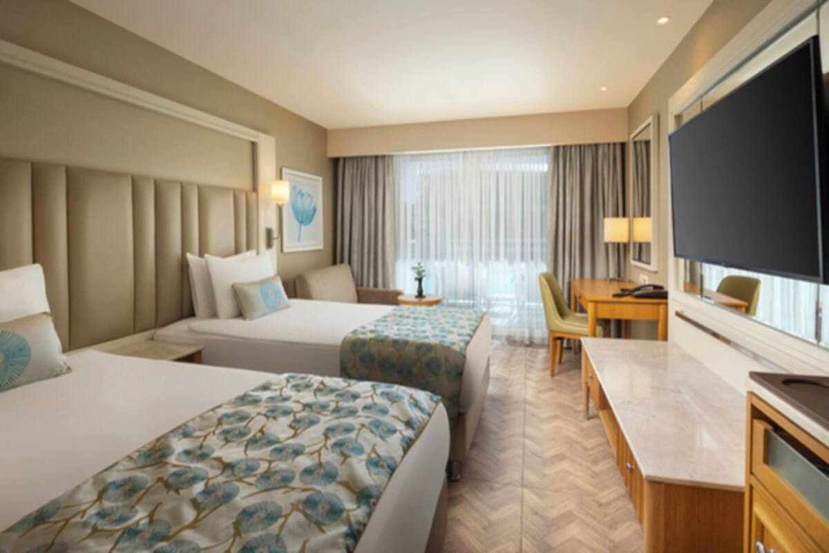 Quarto de hotel com duas camas, uma escrivaninha, televisão e uma janela grande com cortinas.