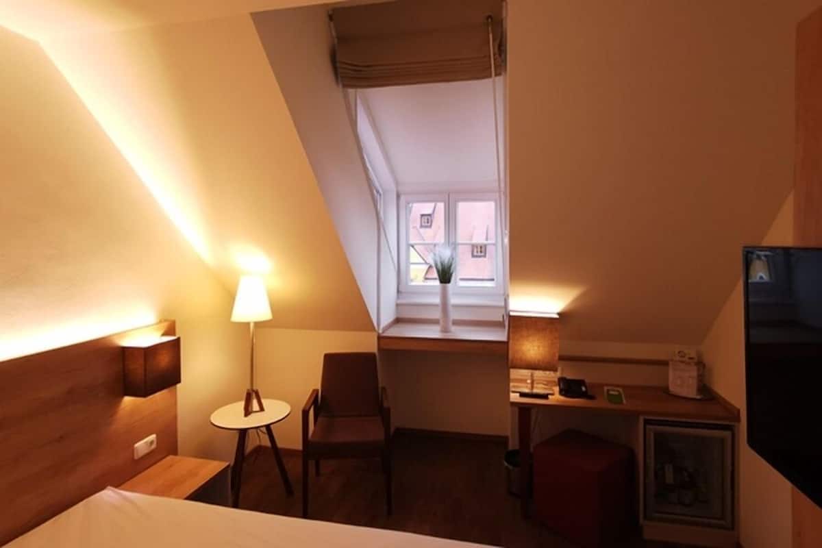 Habitación de hotel con cama, escritorio, silla, mesita, lámpara, televisor y ventana con vistas.