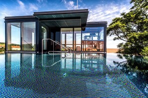 A modern building with a glass exterior, a pool with a reflective surface, and a view of a tree and a distant structure.
