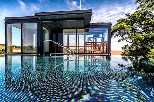 A modern building with a glass exterior, a pool with a reflective surface, and a view of a tree and a distant structure.