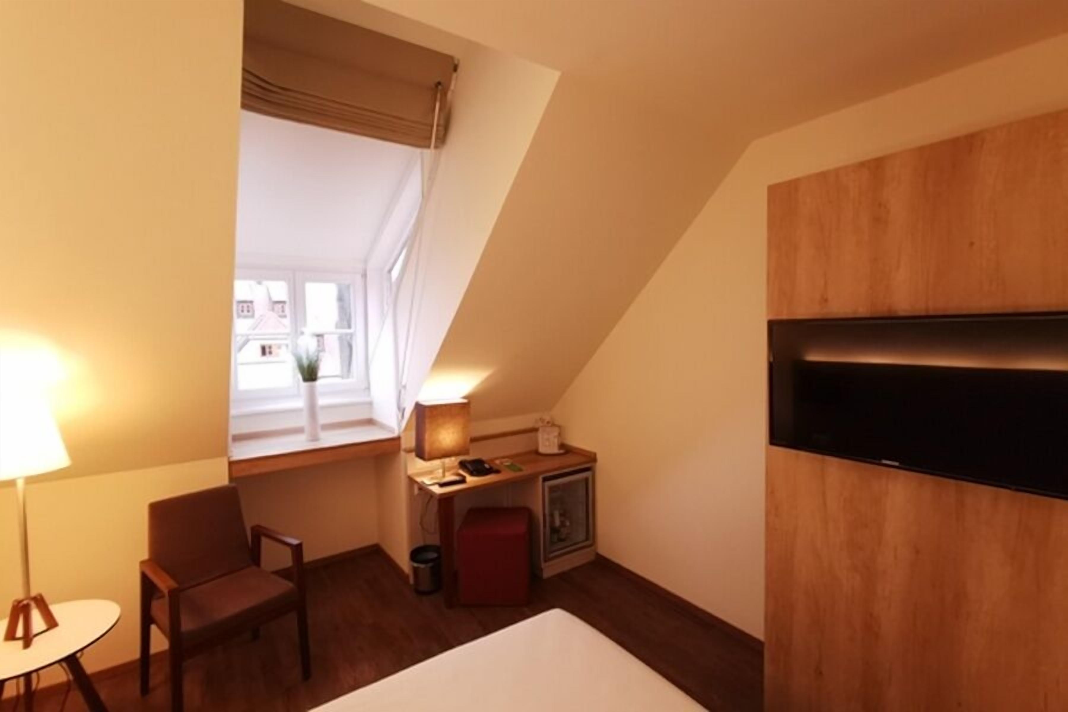 A small, cozy room with a bed, a desk, a chair, a lamp, and a TV mounted on the wall.