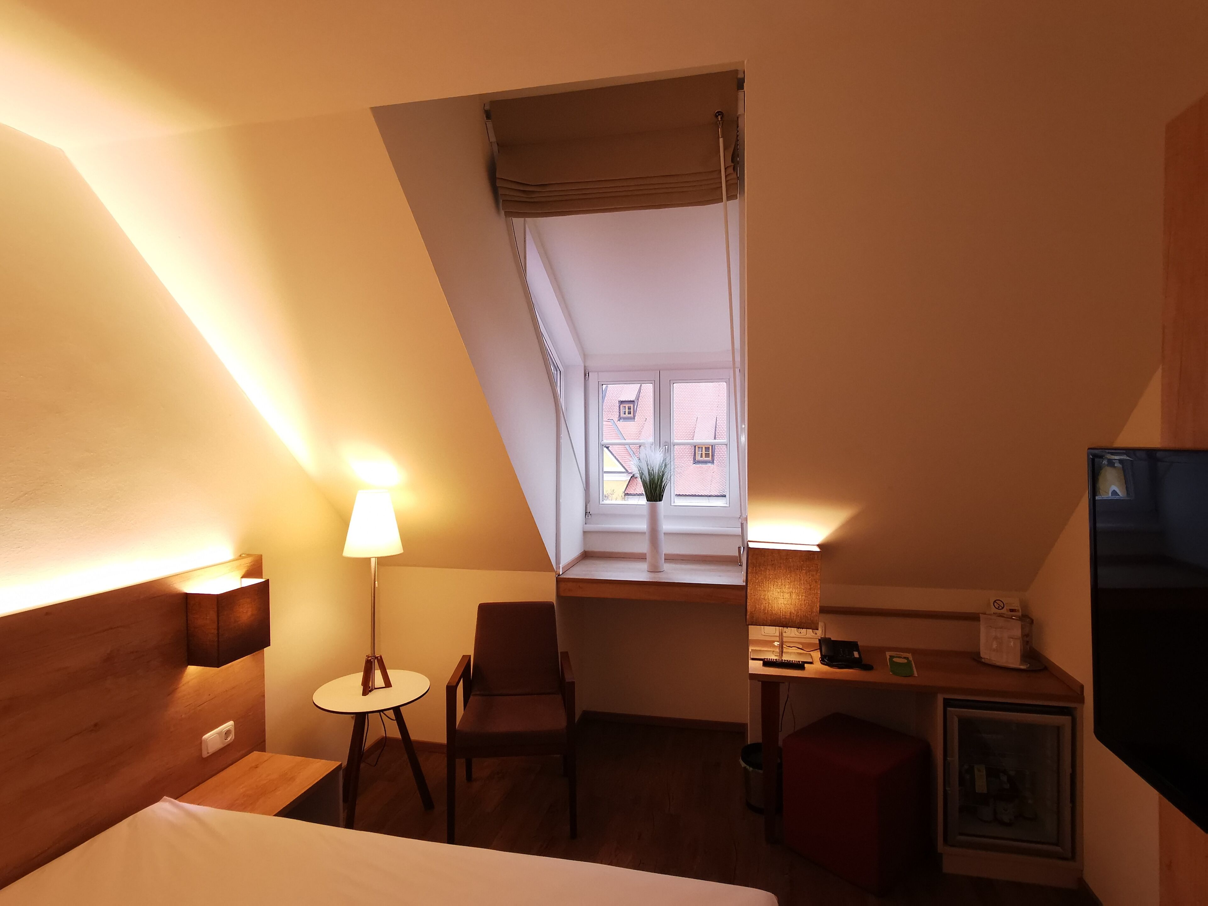 Habitación de hotel con una cama, un escritorio con silla, una mesita, un televisor y una ventana con vistas a edificios.