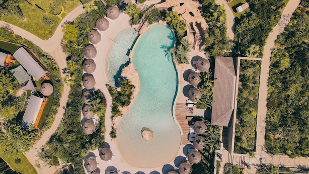 An aerial view of a resort with a central swimming pool, surrounded by beach umbrellas and lounge chairs, with pathways and greenery.