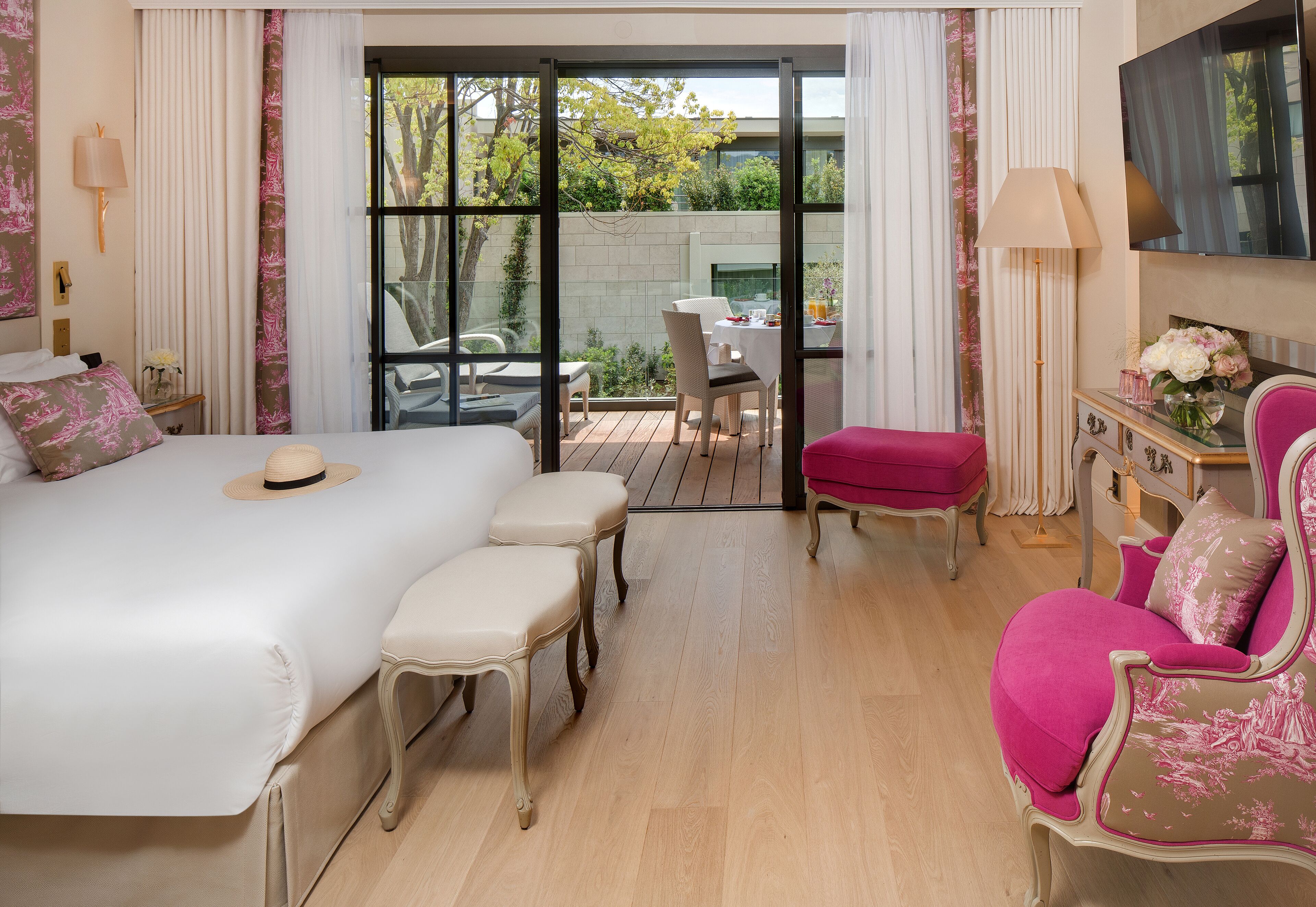 A hotel room with a large bed, a pink armchair, a small table with a vase, and a view of the outdoor area through a sliding door.