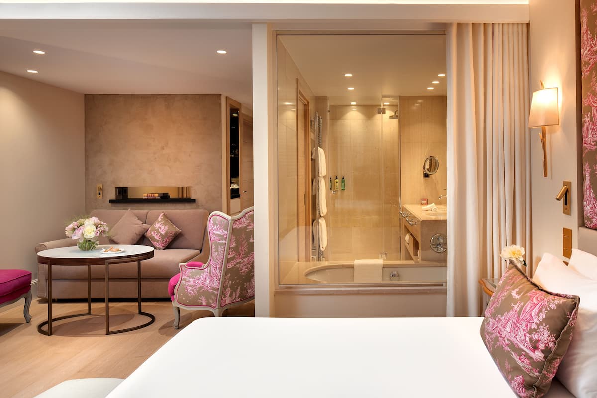 A modern hotel room with a large bed, a round table with a vase of flowers, a sofa, a chair, and a walk-in shower with a glass door.