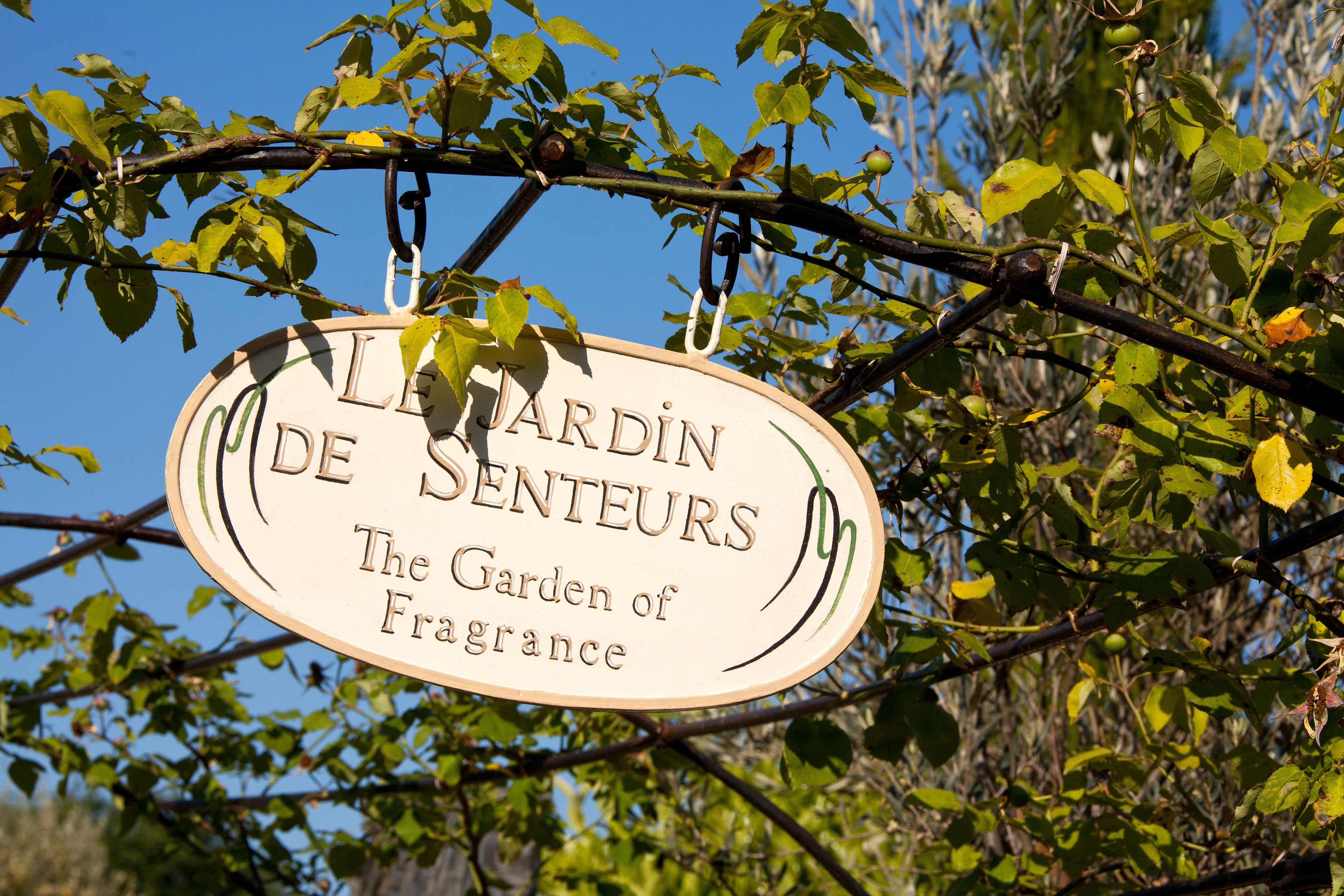 Een bord dat aan een tak hangt, met de tekst 'Le Jardin De Senteurs The Garden of Fragrance'.