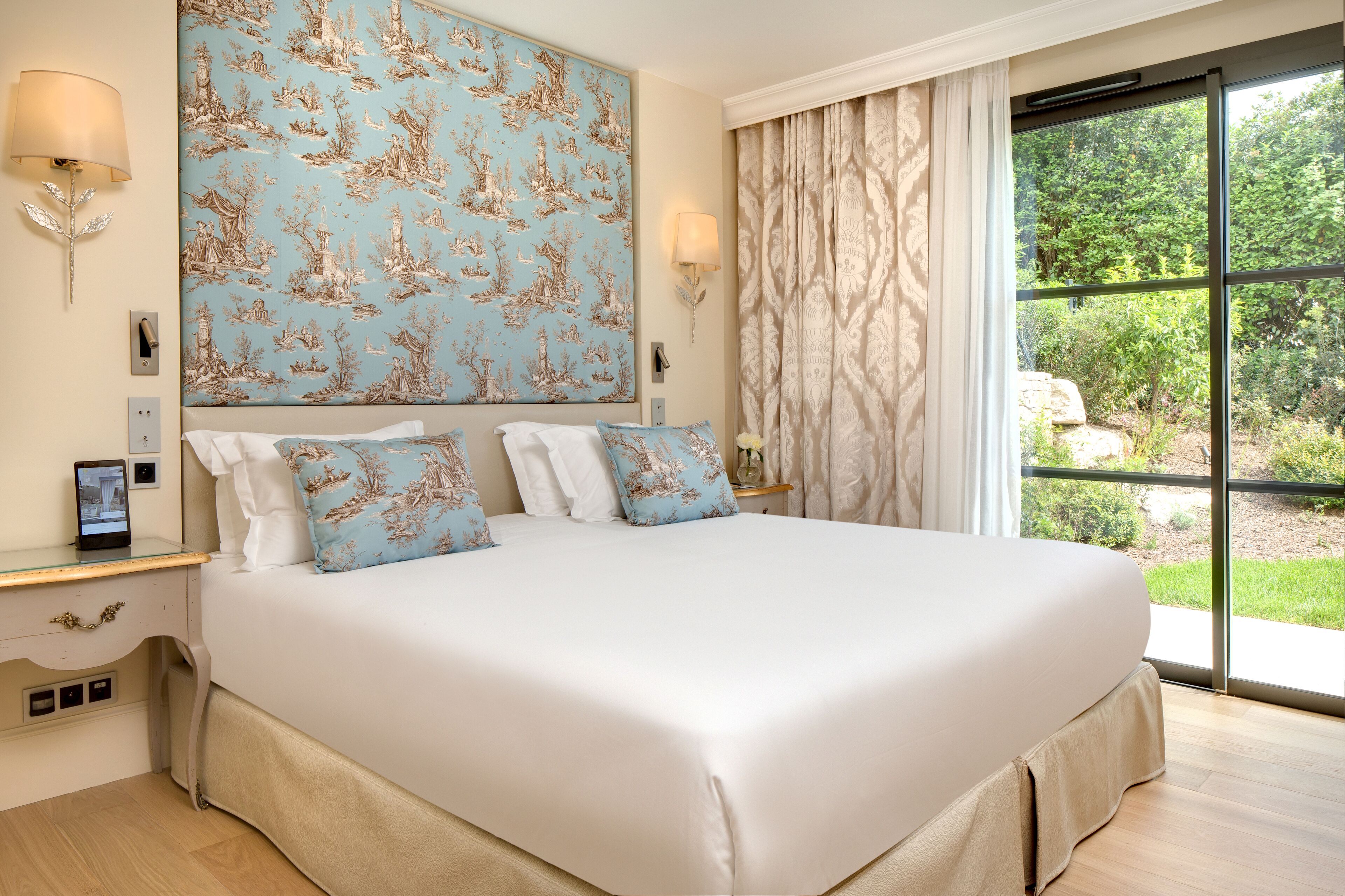 A large bed with white linens and blue patterned pillows, a wall-mounted lamp, and a view of greenery through a large window.