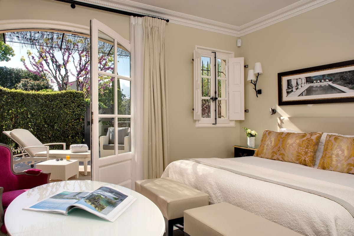 A hotel room with a bed, a small table with a magazine, a chair, a window with curtains, and a wall-mounted lamp.