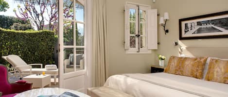 A hotel room with a bed, a small table with a magazine, a chair, a window with curtains, and a wall-mounted lamp.