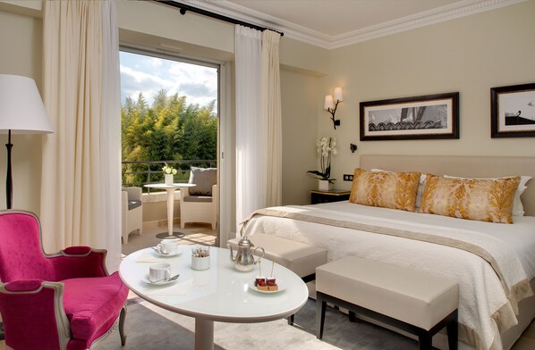 Habitación de hotel con una cama grande, zona de comedor con mesa y sillas, y balcón con vistas a árboles.