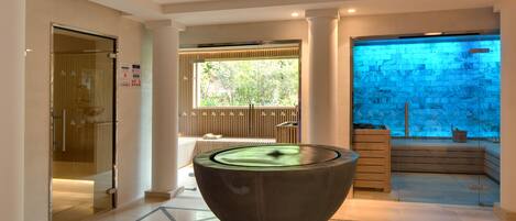 A modern interior with a large, circular water feature in the center, a glass enclosure with a view of greenery outside, and a wooden bench by the window.