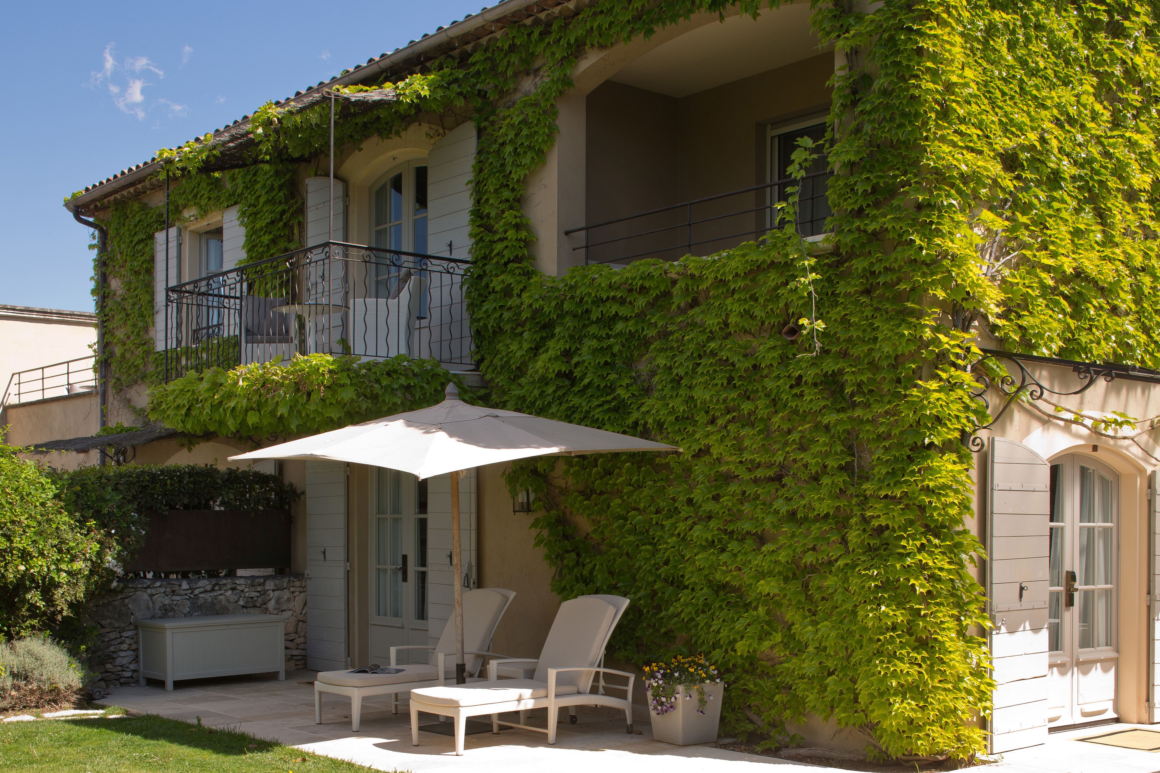A two-story building with a balcony covered in green vines, outdoor seating with a table and chairs, and a large umbrella providing shade.