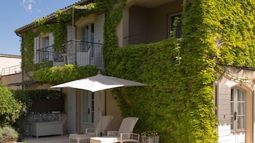 A two-story building with a balcony covered in green vines, outdoor seating with a table and chairs, and a large umbrella providing shade.