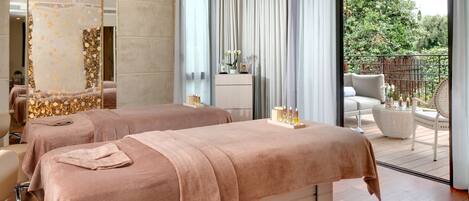 A spa room with two massage tables, a large mirror, and a view of the garden through a sliding door.
