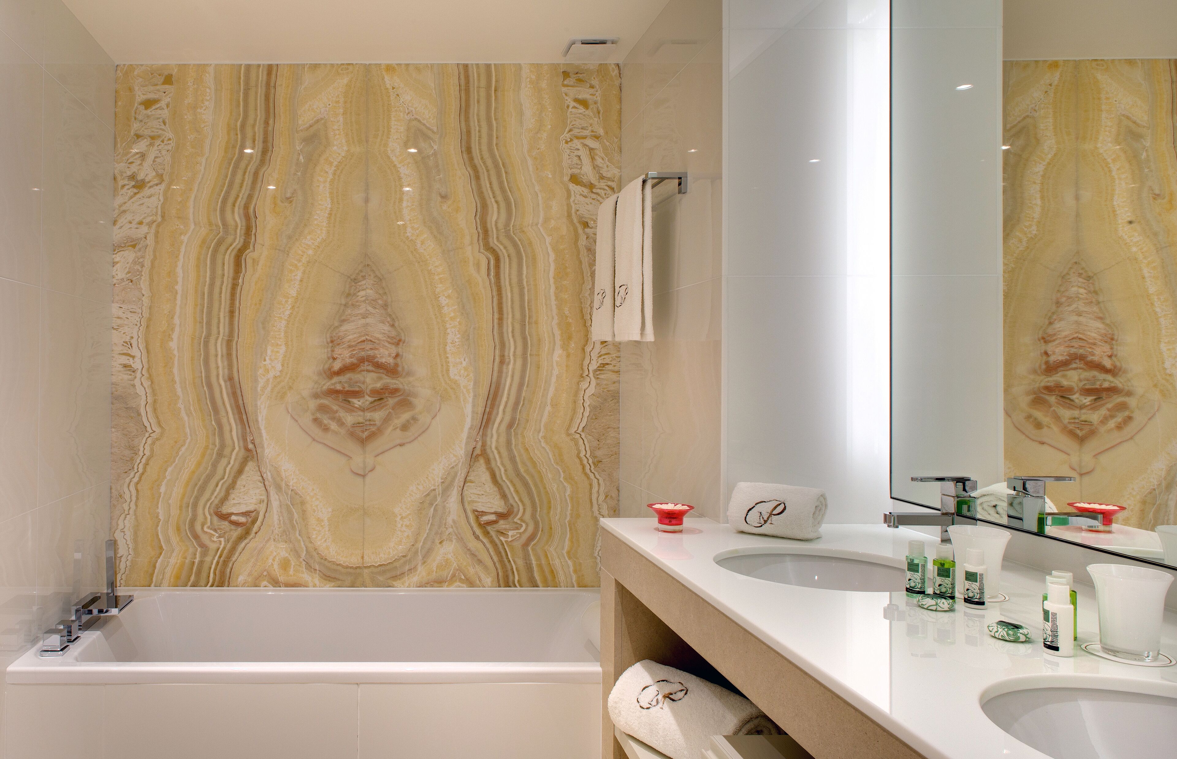 Un baño moderno con pared de mármol, bañera, lavamanos con artículos de aseo personal y espejo.