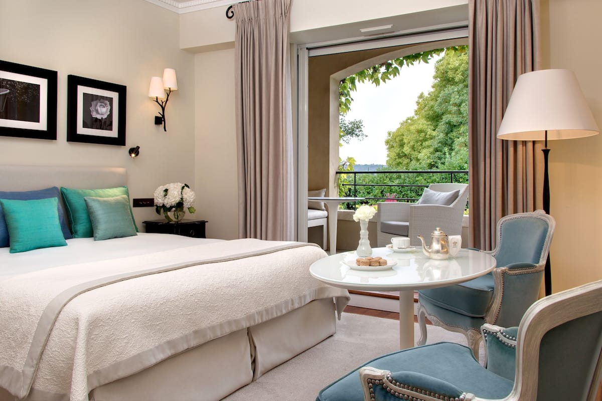 A hotel room with a bed, a round table with a lamp, and a balcony with outdoor seating.