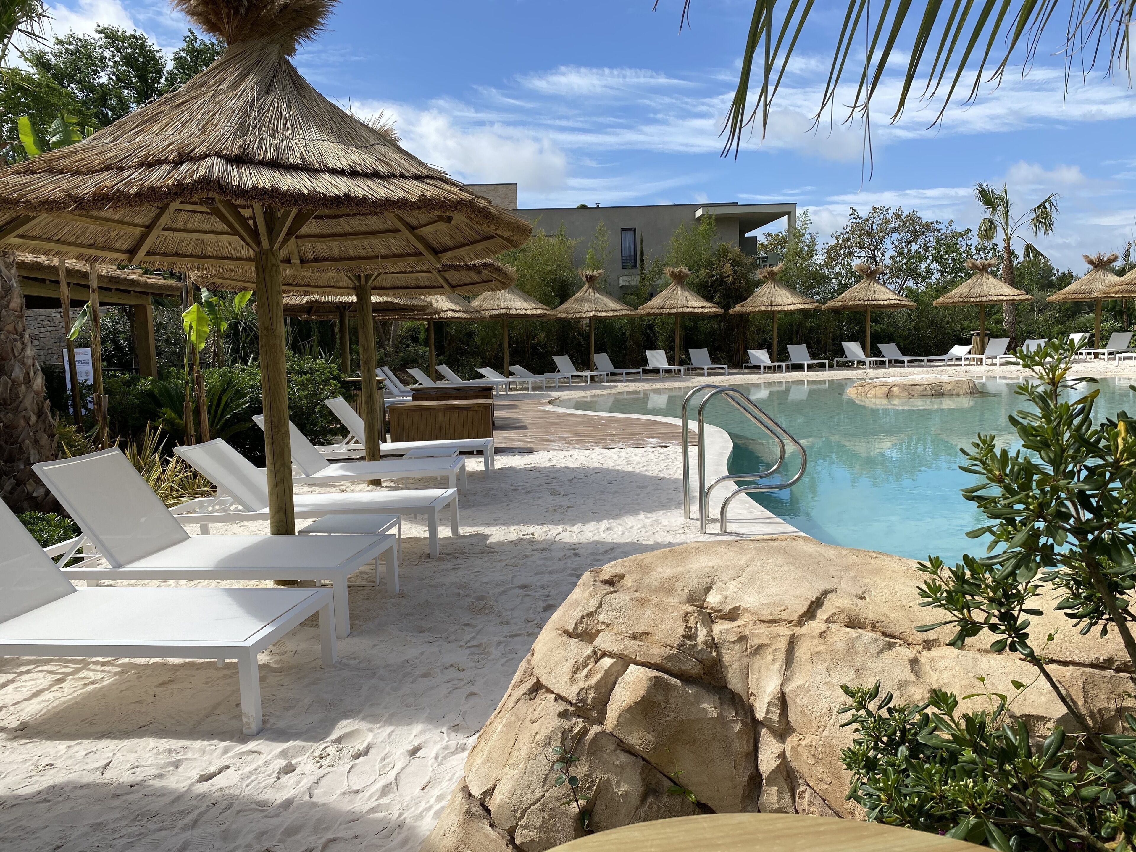A poolside area with white lounge chairs and thatched umbrellas, surrounded by greenery and a clear sky.