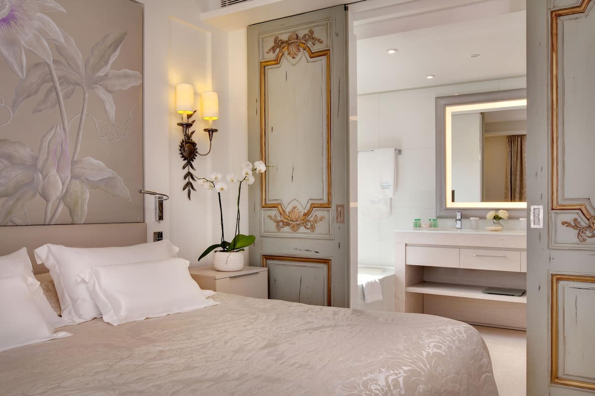A bedroom with a large bed, a nightstand with a lamp, a wall-mounted mirror, and a door leading to a bathroom.