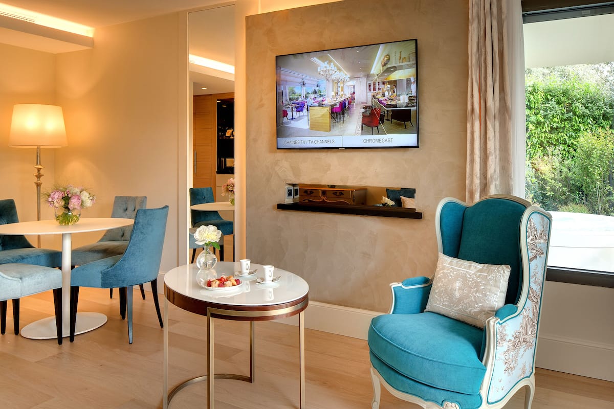 A hotel room with a dining area, a TV mounted on the wall, and a window with a view of greenery.
