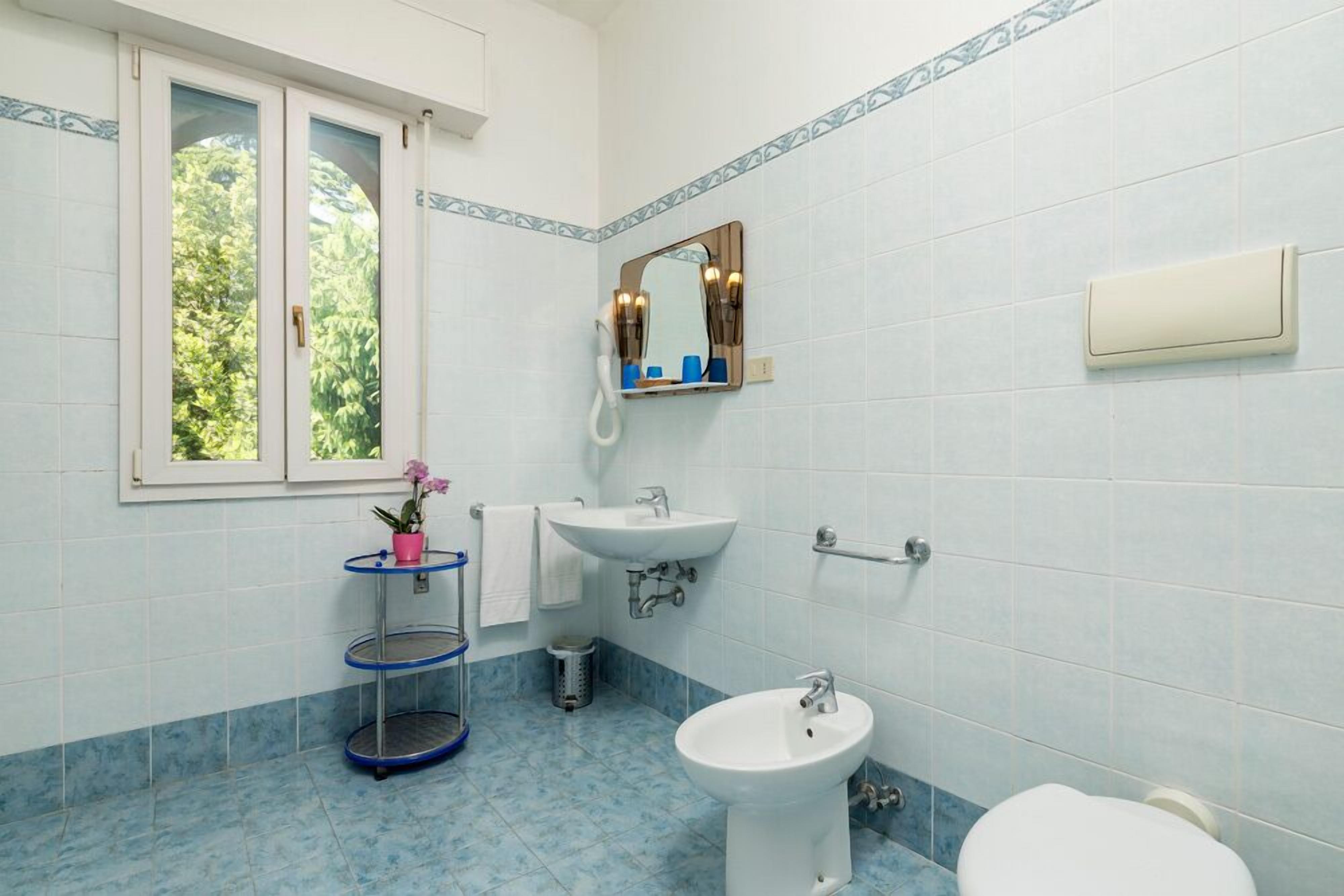 A bathroom with a sink, bidet, and toilet. There is a window with a view of greenery outside.