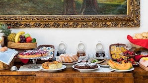 A buffet table with various food items, including fruits, pastries, and desserts, arranged on a wooden table with a painting in the background.