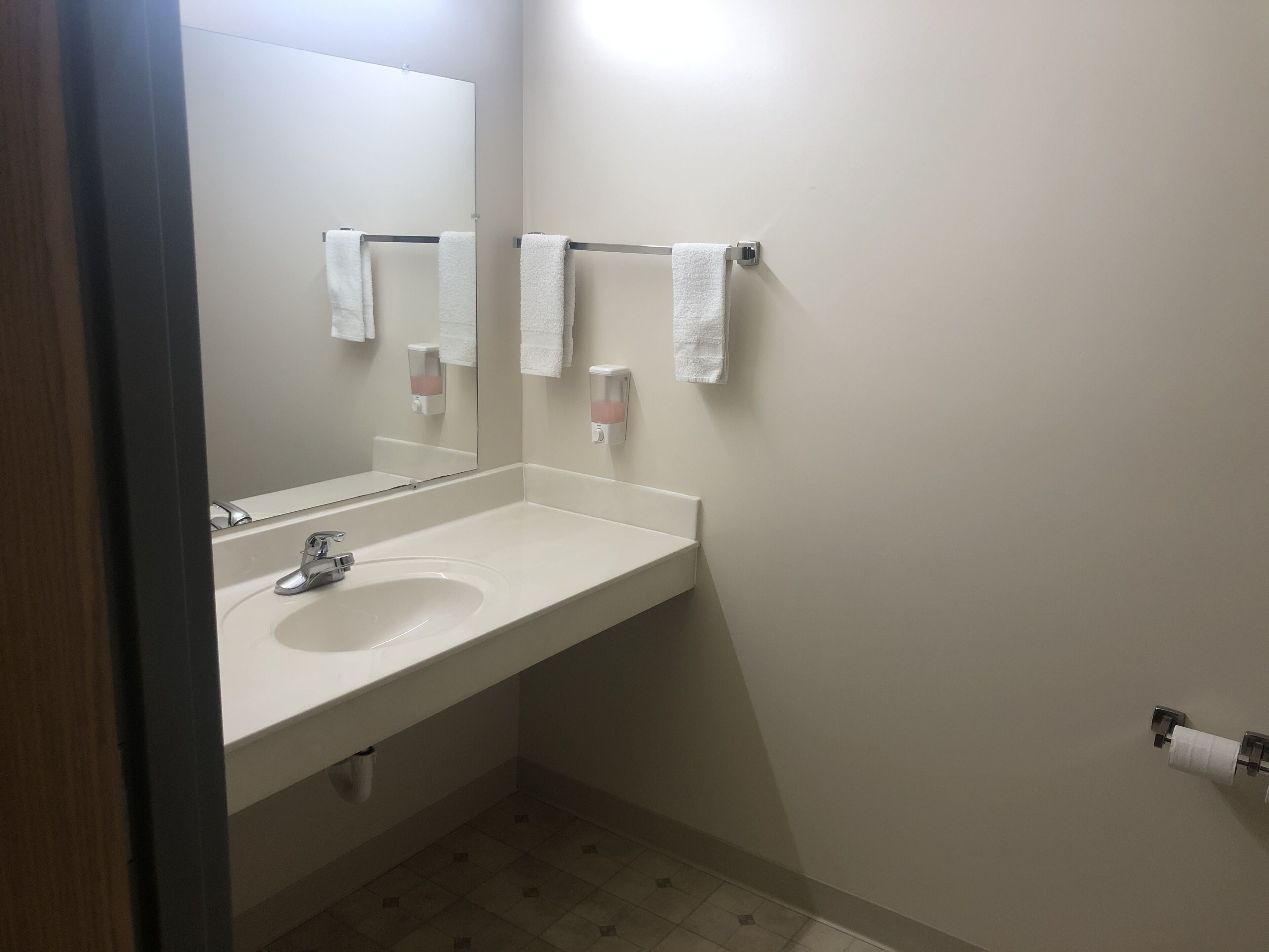 Une salle de bain équipée d’un lavabo, d’un miroir, d’un porte-serviettes, d’un distributeur de savon et de papier toilette.