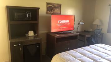 A hotel room with a bed, a TV displaying the word "roman," a microwave, a small refrigerator, a desk with a lamp, and a painting on the wall.