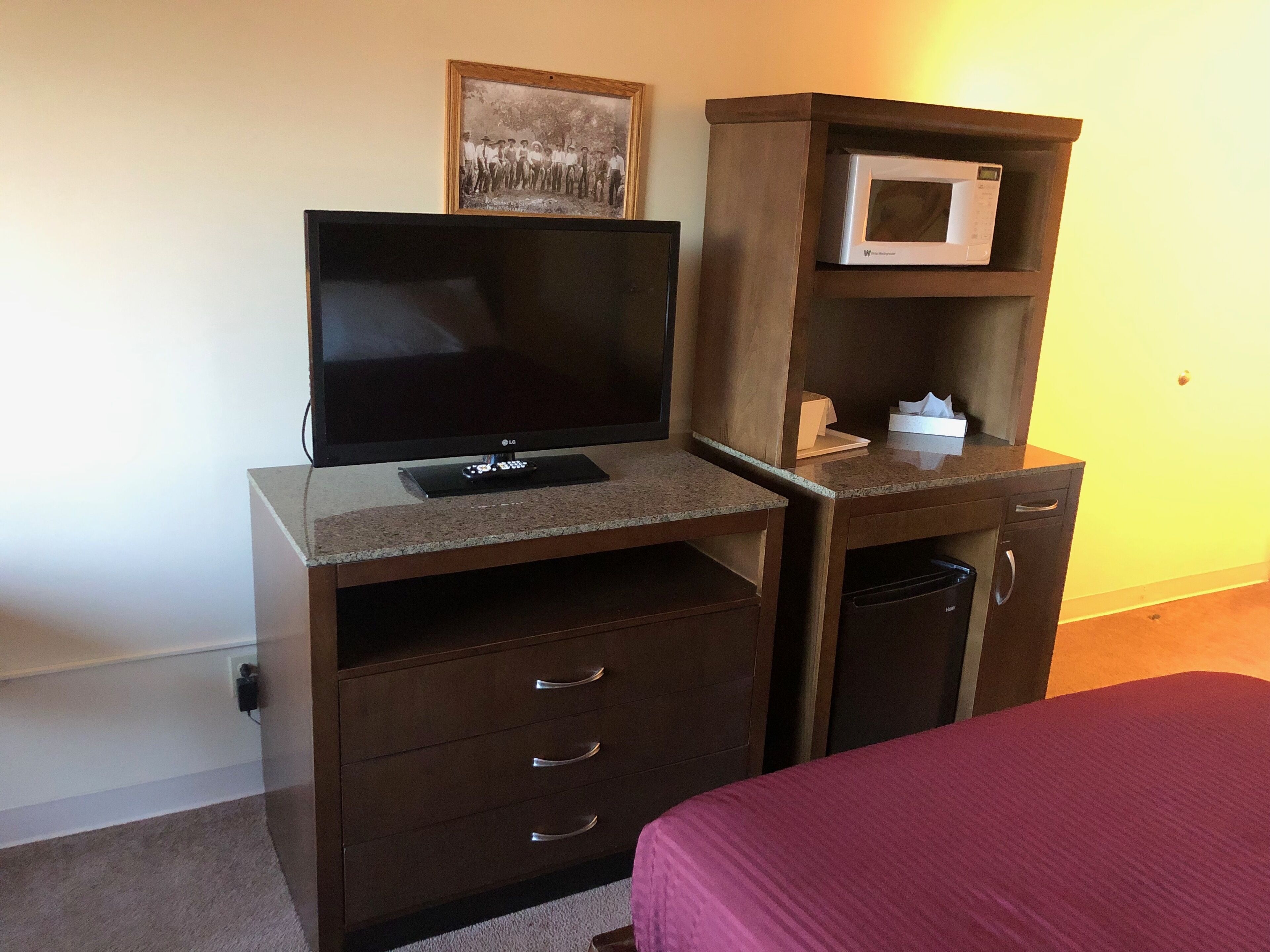 A hotel room with a TV on a wooden desk, a microwave, a small cabinet, and a bed with a red bedspread.
