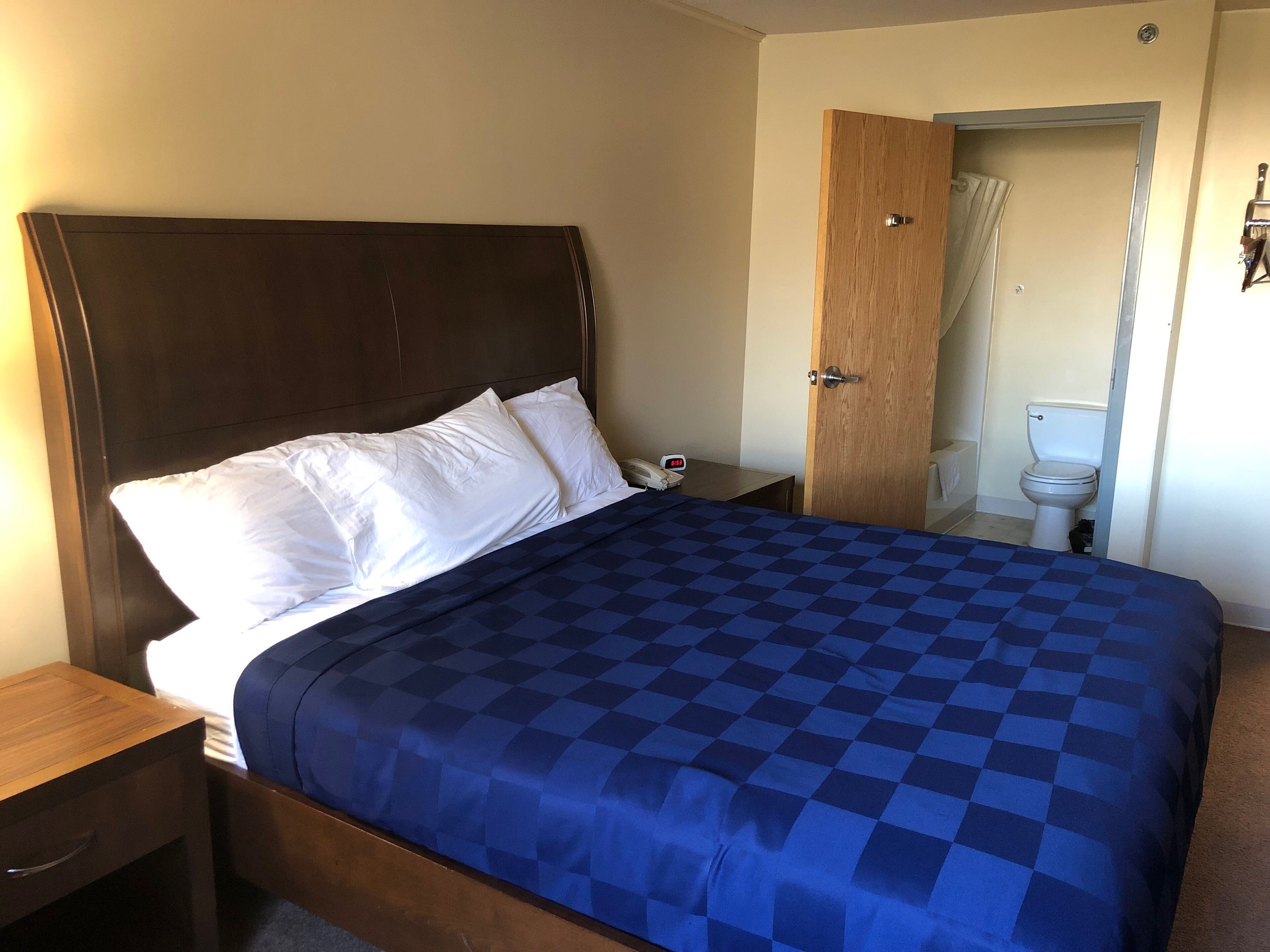 Una cama bien hecha con colcha azul a cuadros y almohadas blancas, una mesita de noche de madera con un teléfono, y una puerta de madera que da a un baño con inodoro y cortina de ducha.