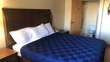 Una cama bien hecha con colcha azul a cuadros y almohadas blancas, una mesita de noche de madera con un teléfono, y una puerta de madera que da a un baño con inodoro y cortina de ducha.