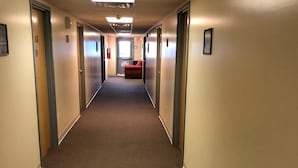 Un long couloir étroit, dont le sol est recouvert de moquette, avec des portes de part et d’autre, et une chaise rouge au loin.