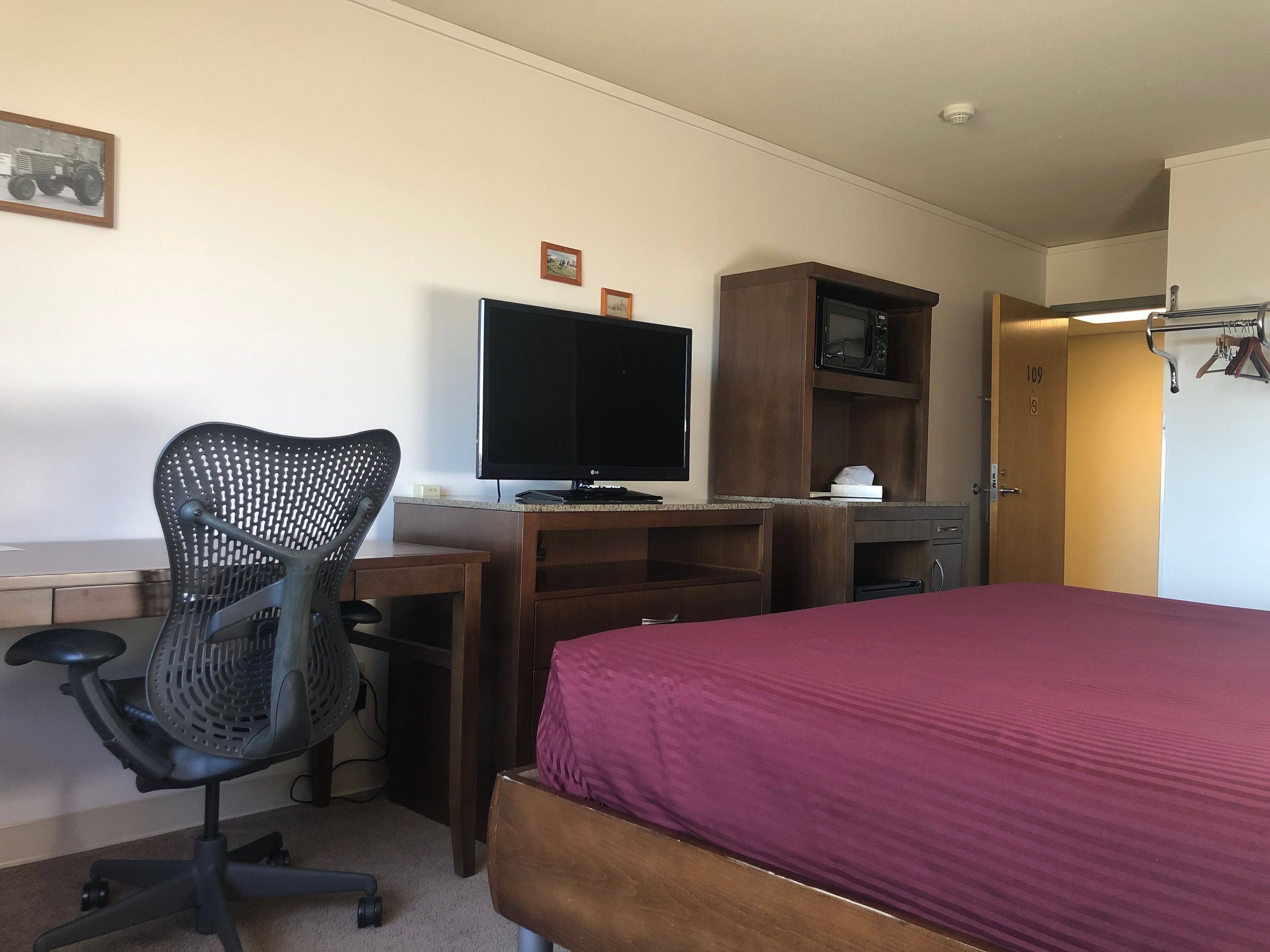 Une chambre d’hôtel comprenant un lit, un bureau avec un ordinateur et une chaise, une télévision, un four à micro-ondes et une porte avec un numéro.
