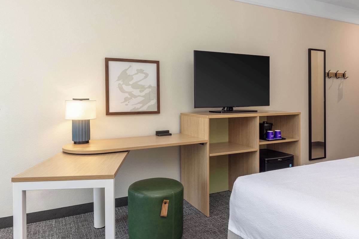 Ein Hotelzimmer mit einem Bett, einem Schreibtisch, einem Fernseher, einer Lampe, einem kleinen grünen Hocker, einem Regal mit Tassen und einem Spiegel.