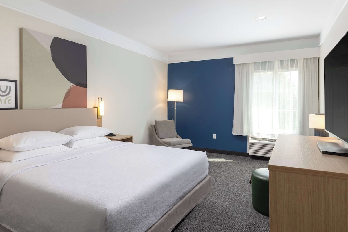 Ein Hotelzimmer mit einem großen Bett, einem Schreibtisch, einem Stuhl, einer Lampe und einem großen, abstrakten Gemälde an der Wand.