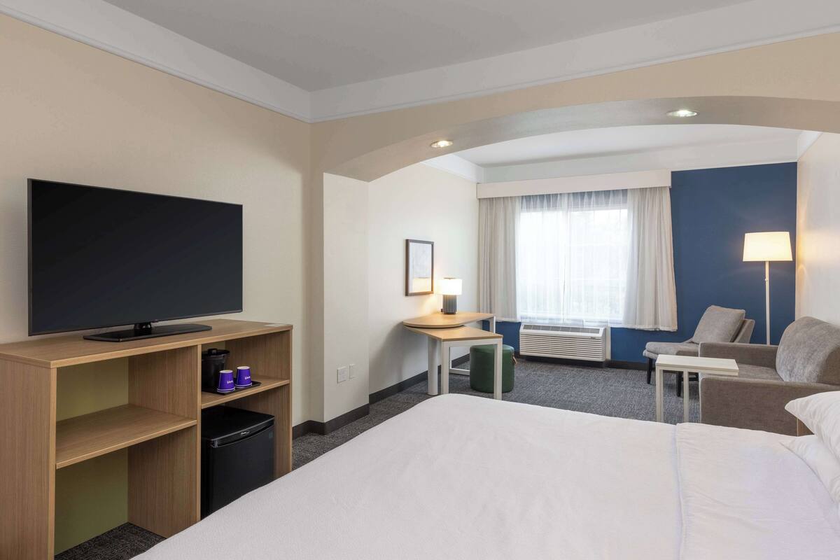 Ein Hotelzimmer mit einem großen Bett, einem Fernseher auf einer Holzeinrichtung, einem Schreibtisch mit Lampe, einem Stuhl und einem Fenster mit Vorhängen.