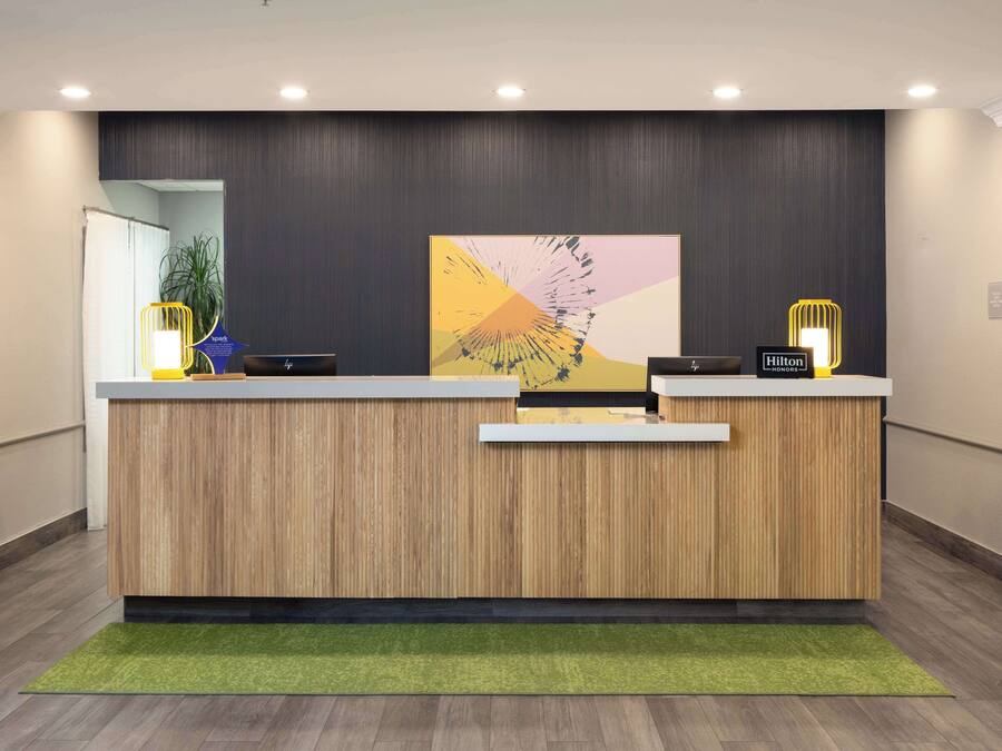 Un comptoir d’accueil au design moderne, avec un revêtement en bois et un tapis vert. Deux panneaux lumineux sont disposés sur le comptoir, l’un indiquant « Hilton Honors » et l’autre le nom de l’hôtel.