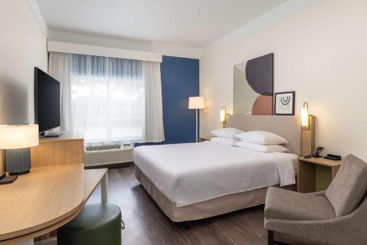 A hotel room with a bed, a desk, a chair, a TV, a lamp, and a window with curtains.
