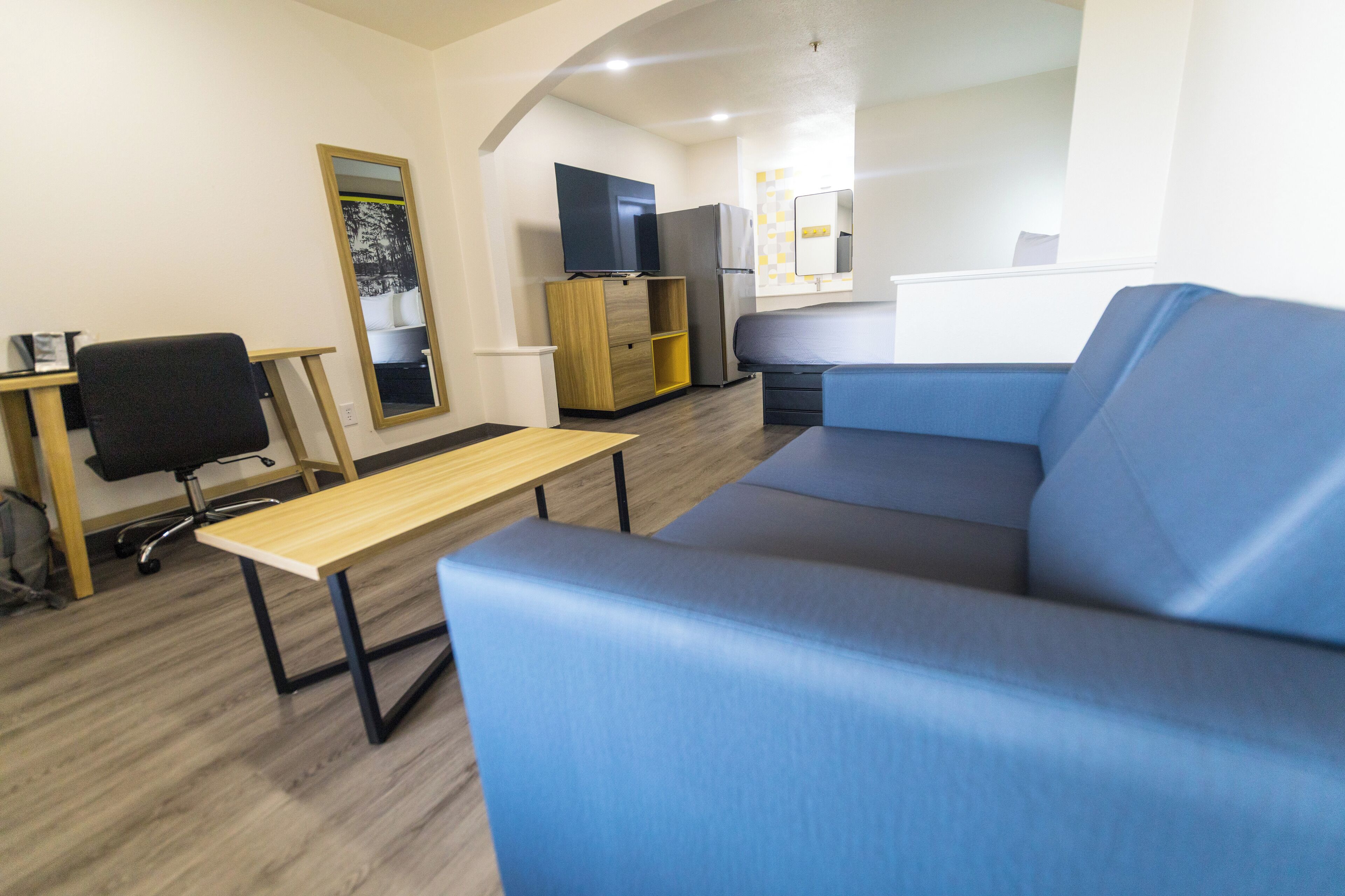 A modern living room with a blue sofa, a wooden coffee table, a black office chair, and a TV on a wooden cabinet.