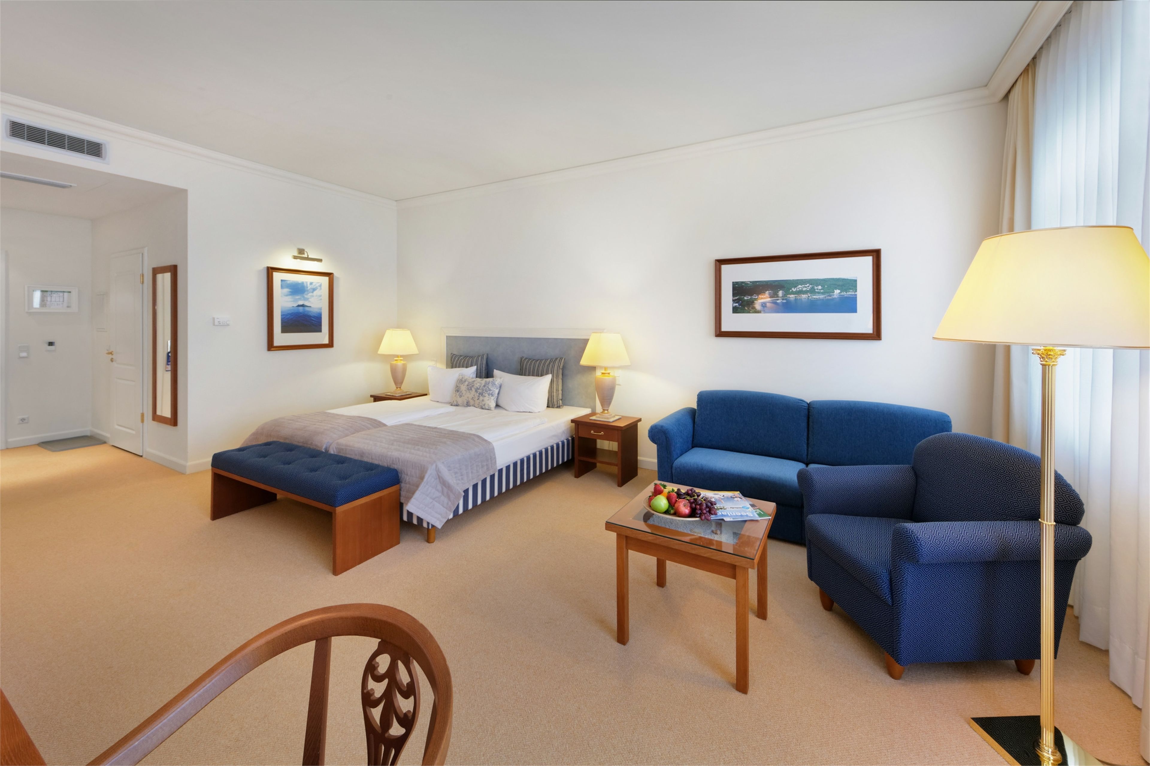 Quarto de hotel com uma cama, um sofá azul, uma pequena mesa com frutas e um abajur.