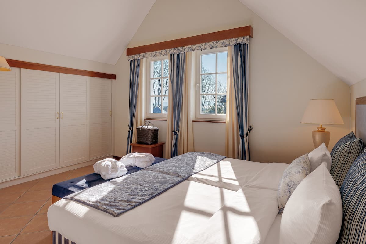 A bedroom with a bed, a nightstand, a lamp, a window with curtains, and a wardrobe.