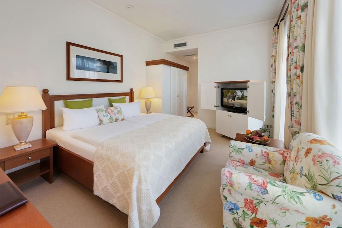 Una habitación de hotel con una cama, dos mesitas de noche con lámparas, un sillón tapizado con motivos florales, un televisor y un armario.