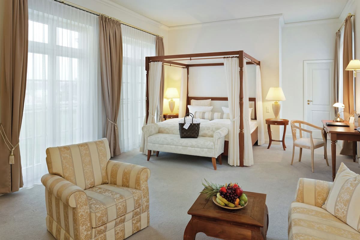 A four-poster bed with a canopy, a sitting area with a sofa and armchairs, a wooden coffee table with fruit, and a desk with lamps.