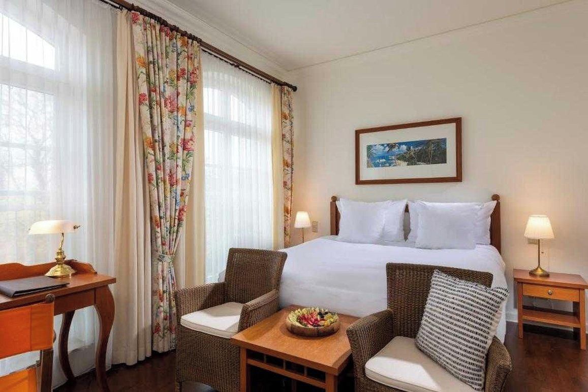 A hotel room with a bed, two chairs, a small table with a fruit bowl, a desk with a lamp, and a framed picture on the wall.