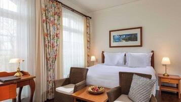 A hotel room with a bed, two chairs, a small table with a fruit bowl, a desk with a lamp, and a framed picture on the wall.