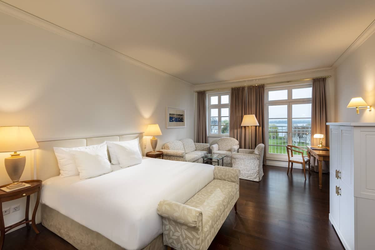 A hotel room with a large bed, two armchairs, a small table, and a view of the outdoors.