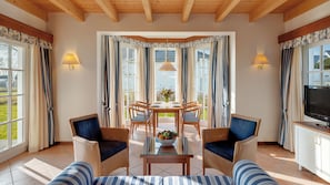 Un comedor con una mesa dispuesta para cuatro personas, rodeada de sillas de mimbre con cojines, un televisor sobre un mueble bajo y grandes ventanales con cortinas.