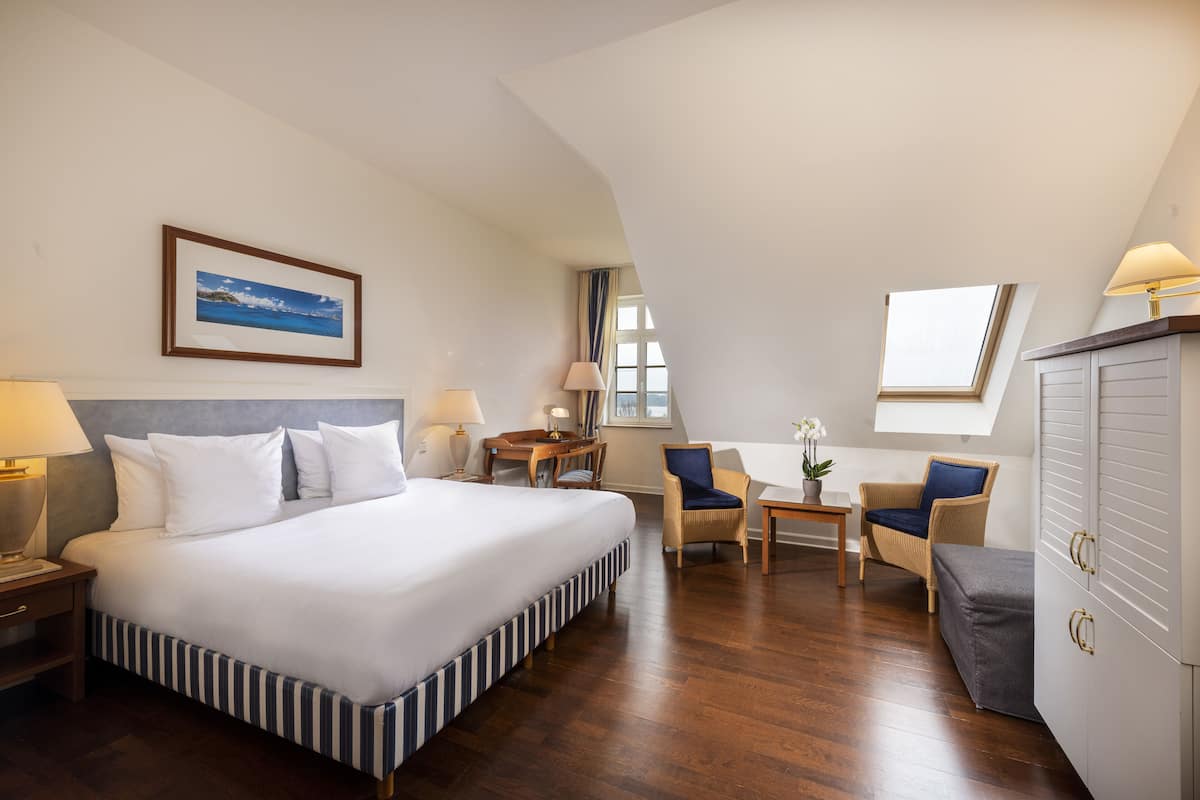 A hotel room with a large bed, a desk, two chairs, a small table with a plant, and a wardrobe.