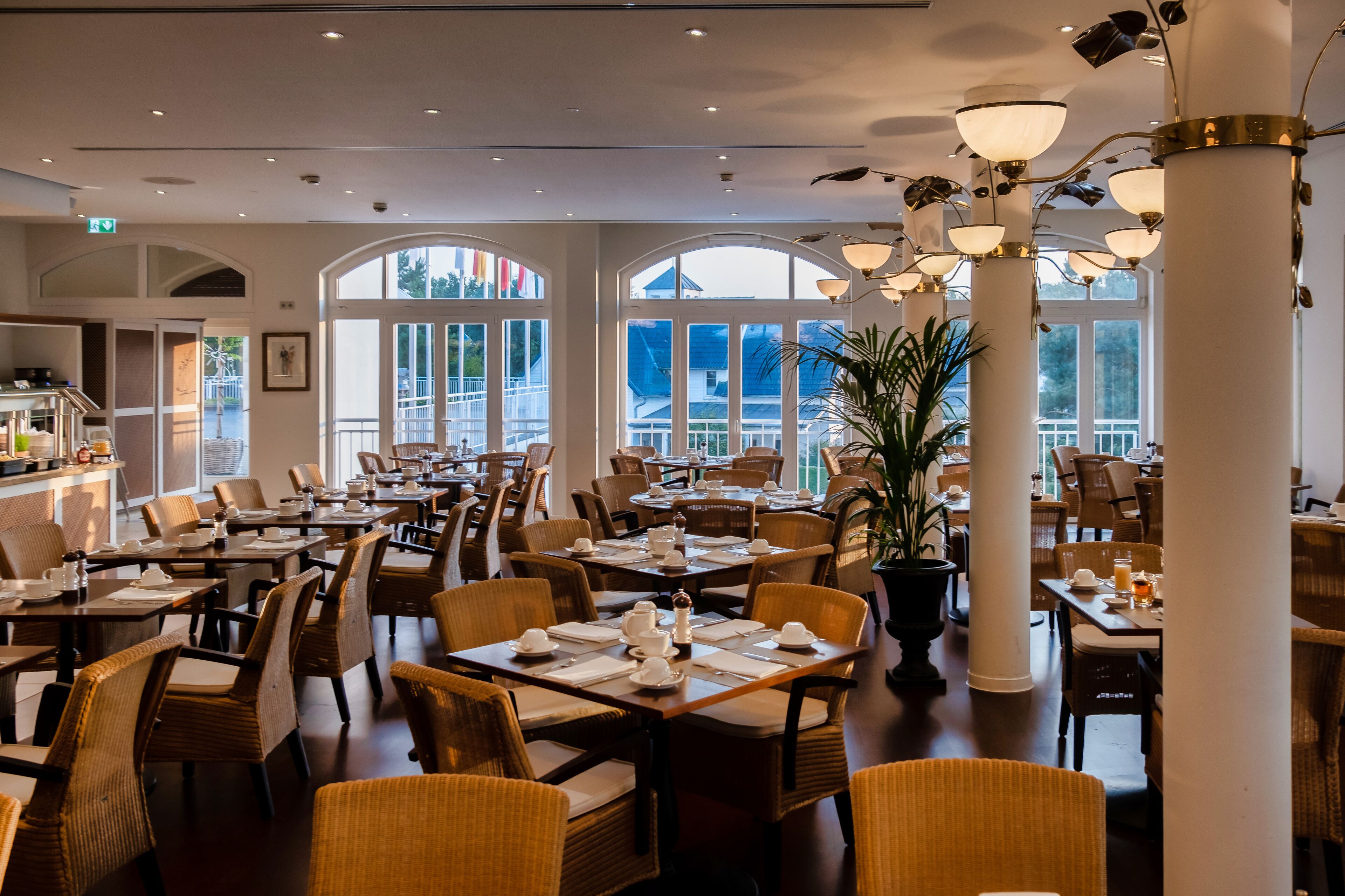 A spacious dining area with high ceilings, large windows, and modern chandeliers. Tables are neatly set with white tablecloths and chairs.
