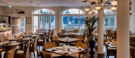Une salle à manger spacieuse avec de hauts plafonds, de grandes fenêtres et des lustres modernes. Les tables sont élégamment dressées avec des nappes blanches et des chaises assorties.